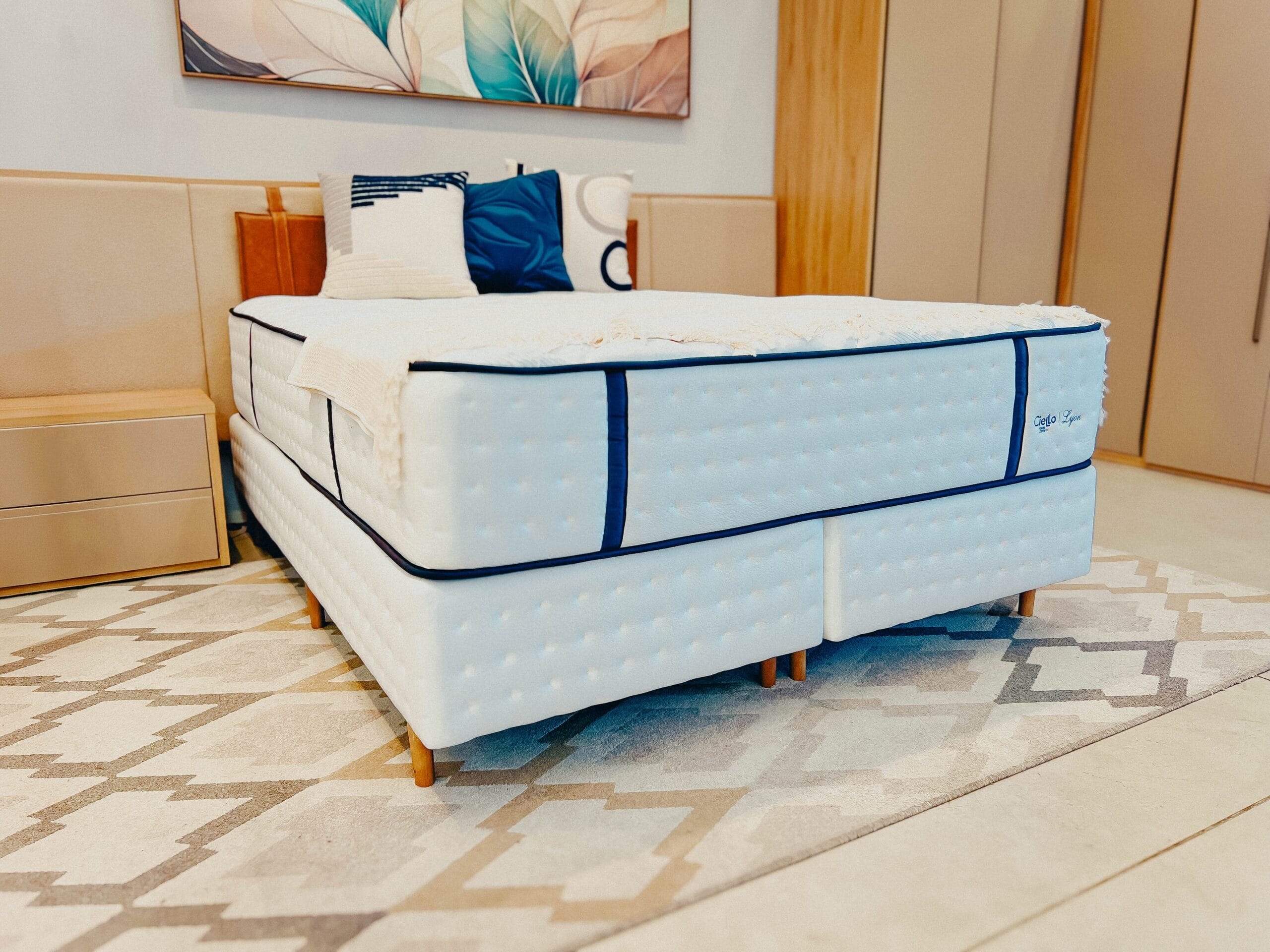 Cama box branca com detalhes azuis e tecnologia Sense Ice em um quarto moderno com tapete geométrico.