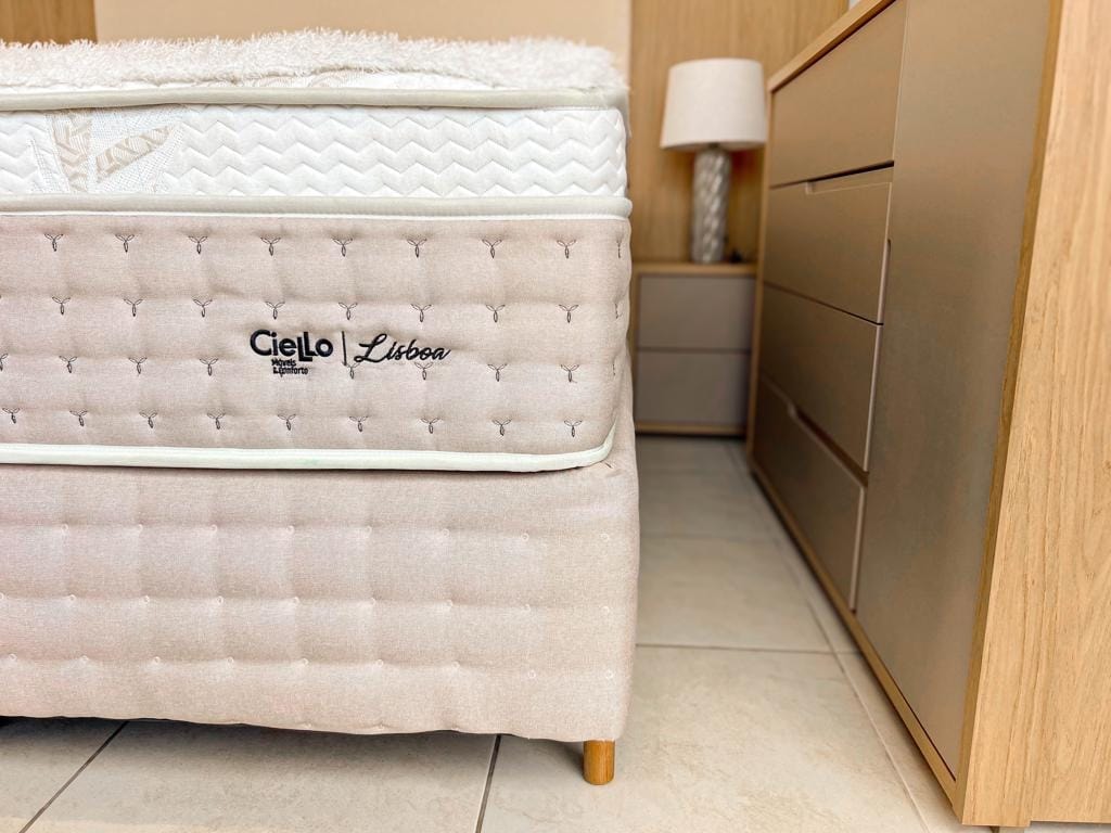 Visão frontal da cama Lisboa arrumada com travesseiros brancos volumosos, destacando a superfície macia e a cabeceira acolchoada.