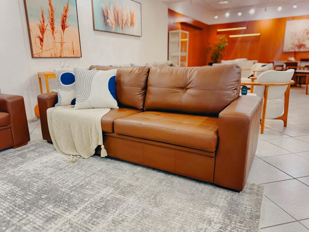 Vista frontal do Sofá Fixo Fit em couro legítimo caramelo com assento em matelassê quadriculado, decorado com almofadas geométricas e manta em um showroom de móveis.