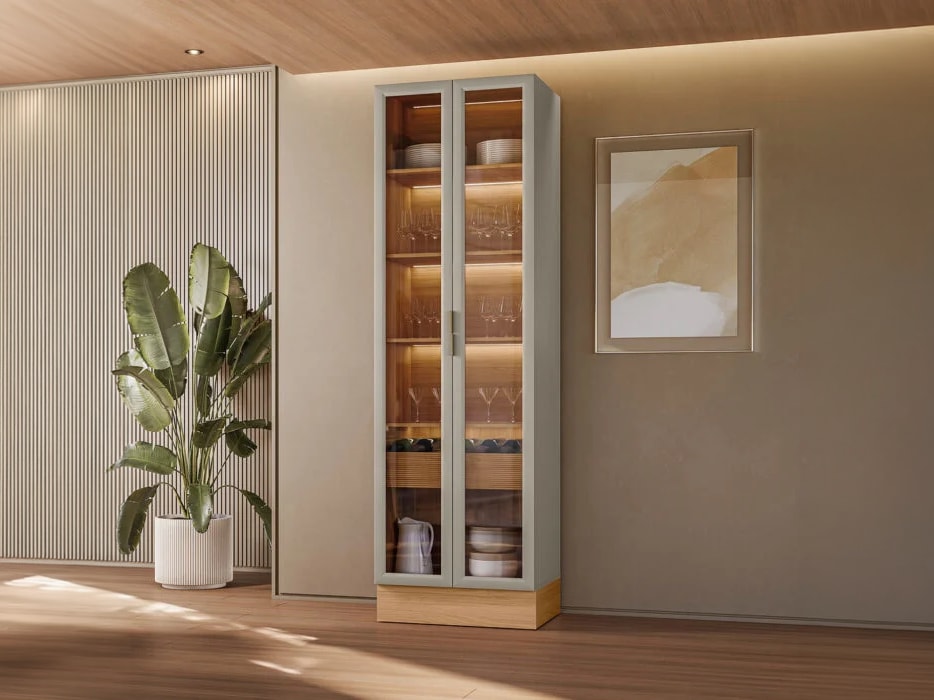 Vista frontal da Cristaleira Allure alta de duas portas de vidro ambientada em uma sala de jantar moderna de parede bege, ao lado de um vaso com planta ornamental.