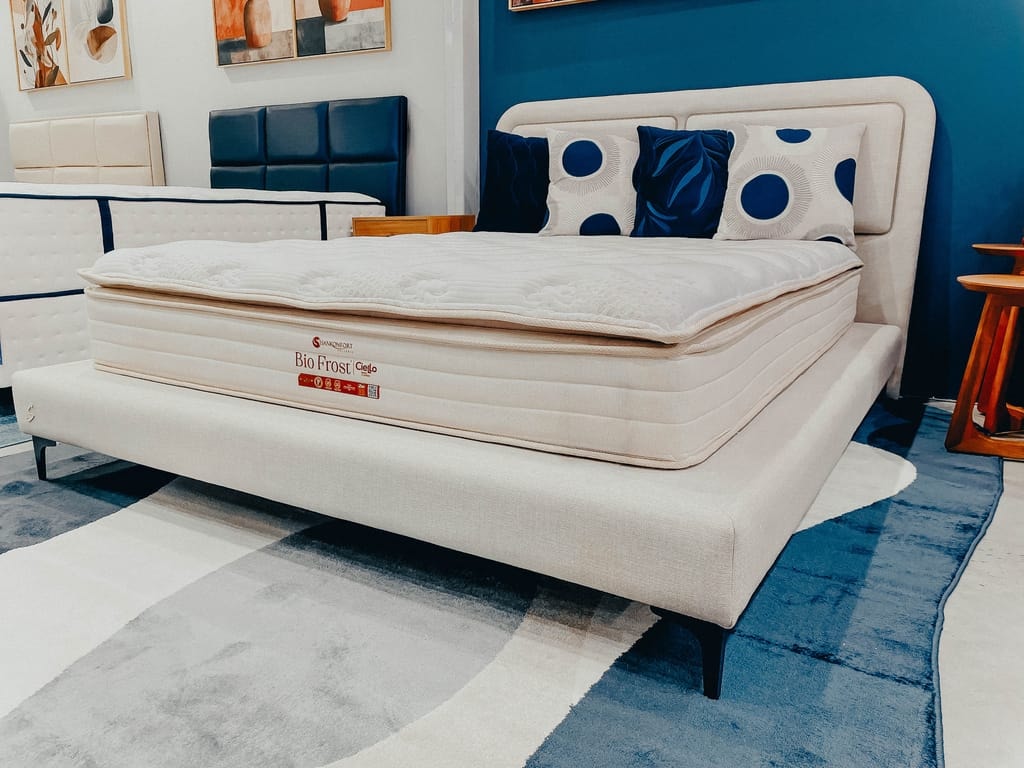 Vista diagonal do colchão Bio Frost de casal sobre uma cama estofada clara, decorada com travesseiros azuis e brancos, contrastando com uma parede azul escura ao fundo.