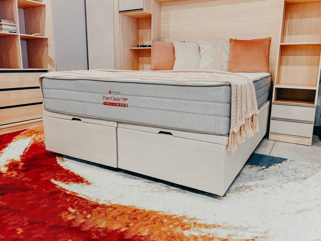 Cama de casal equipada com o colchão Pure Oasis de lateral cinza, apoiado sobre um sommier claro com gavetas frontais fechadas, num quarto decorado com estantes de madeira e tapete laranja.