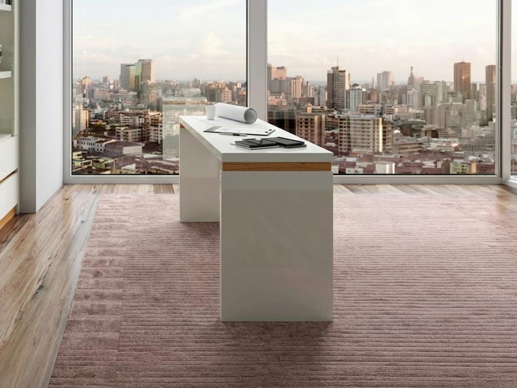 Vista de perfil longitudinal da Mesa de Escritório TC144 branca em MDF, disposta sobre um tapete listrado texturizado em frente a uma grande janela com vista panorâmica para uma grande cidade.