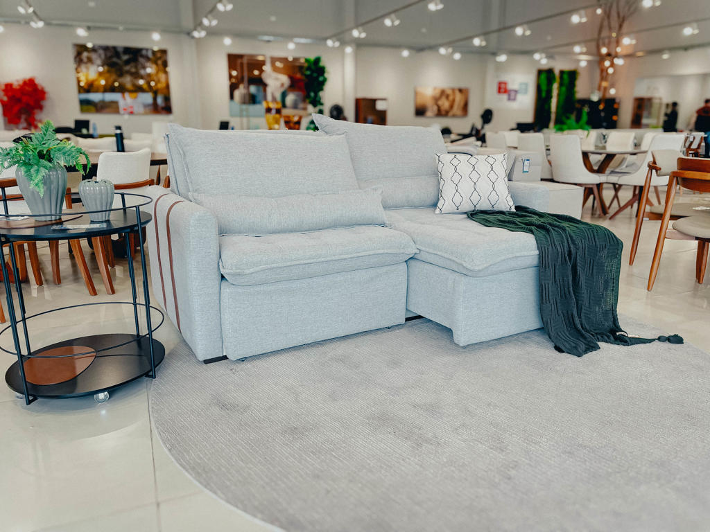 Sofá Retrátil Prestige Cinza Molas Ensacadas Sofá retrátil e reclinável cinza claro em uma sala de exposição, com almofada decorativa e manta verde.