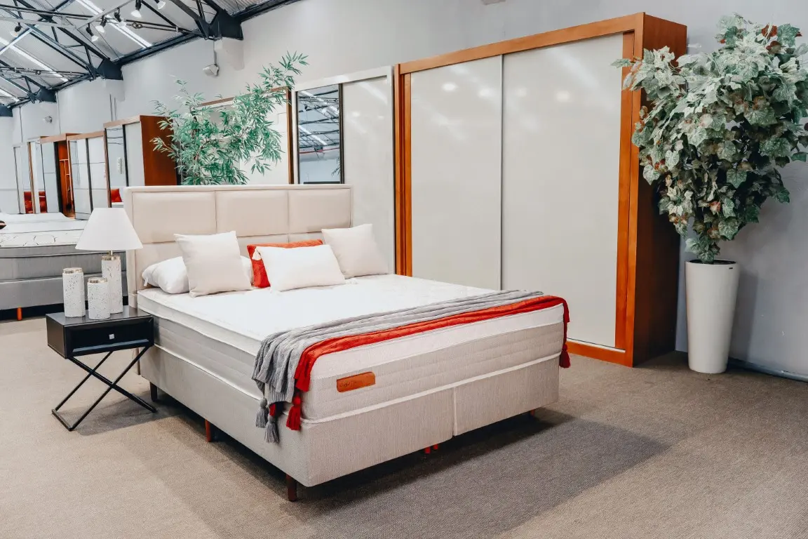 Conjunto cama box Florença em ambiente decorado. Colchão branco alto com tampo em malha Jacquard e base box revestida em tecido linho cinza claro. Detalhe para a manta decorativa laranja e cinza sobre a cama.