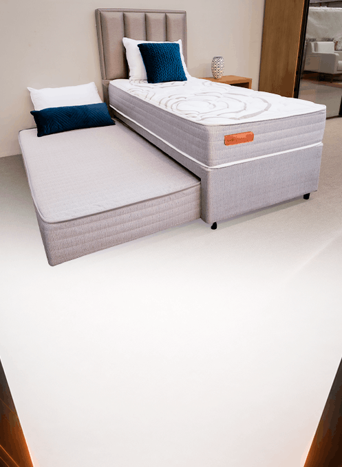 Grey upholstered bed with a matching pull-out trundle in a showroom, white and blue cushions on the bed.