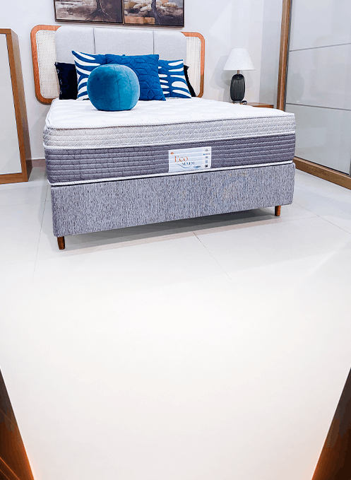 Display bed on show floor with gray upholstered base, blue pillows, and a nearby lamp on a nightstand.