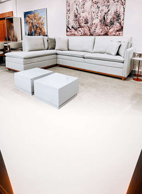 Modern showroom living room: light gray L-shaped sofa, two pale blue coffee tables, and pink floral wall art above the sofa.