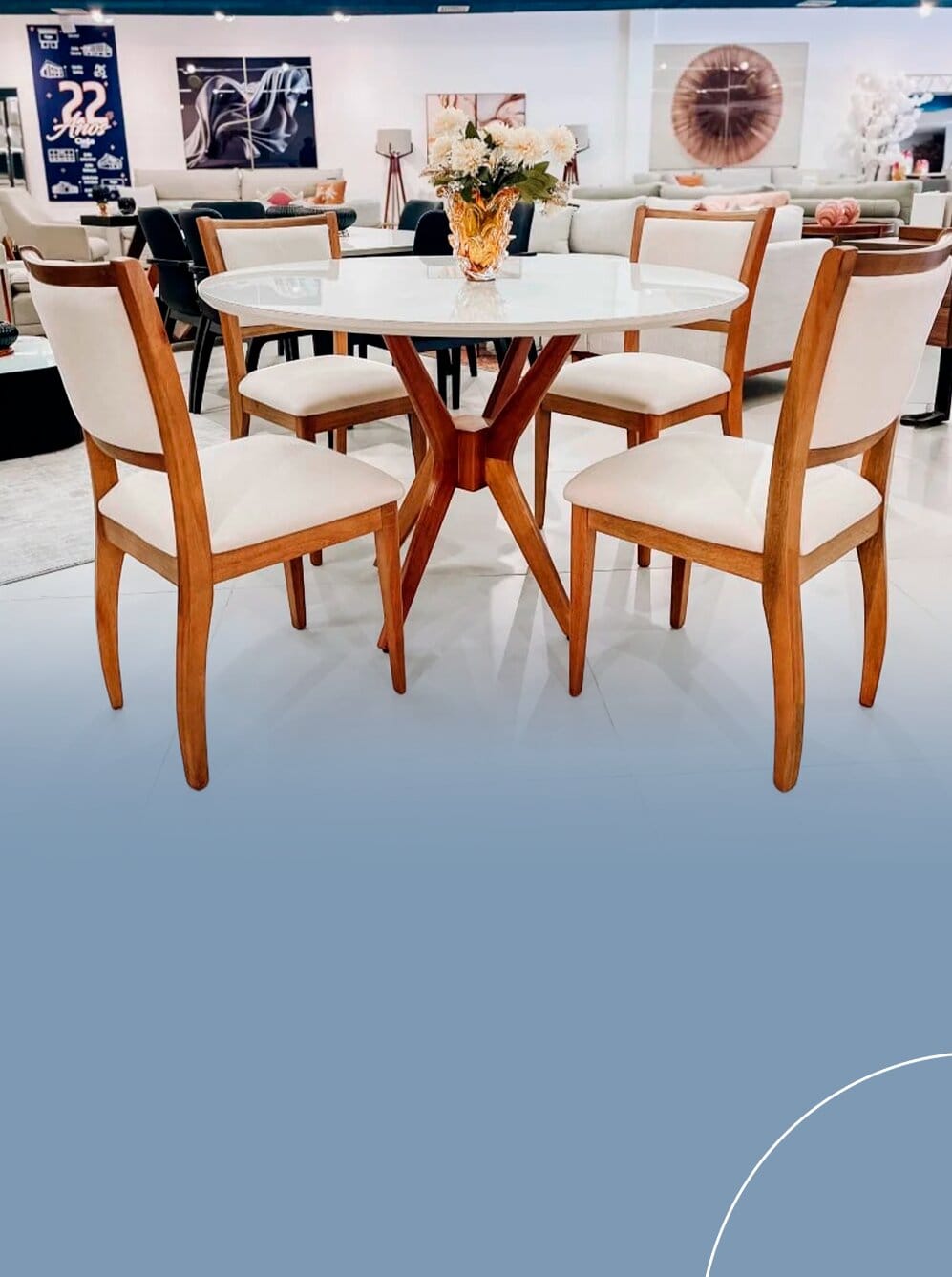 Conjunto de mesa de jantar redonda com tampo branco e base central de madeira cruzada, acompanhada de quatro cadeiras de encosto alto estofadas em tom off-white.