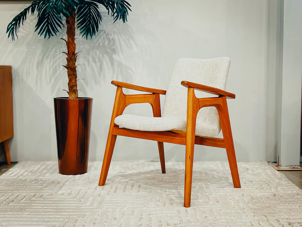 Poltrona Escandinava Madeira Aline | Ciello Visão em perspectiva da Poltrona Aline com estrutura em madeira maciça de Jequitibá e estofamento claro, ambientada ao lado de uma planta ornamental em vaso acobreado sobre um tapete neutro.