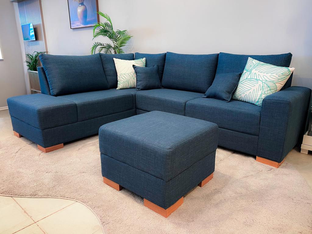 Sofá de Canto Modular Viena Azul | Ciello Visão completa do Sofá de Canto Viena em tecido texturizado azul profundo, composto por um módulo com braço, módulo de canto, extremidade em puff fixo e um puff avulso quadrado no centro da sala.