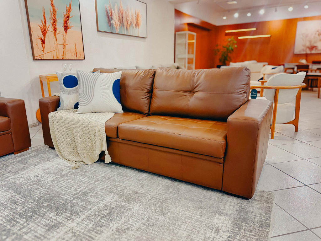 Sofá de Couro Legítimo Caramelo Fit | Ciello Vista frontal do Sofá Fixo Fit em couro legítimo caramelo com assento em matelassê quadriculado, decorado com almofadas geométricas e manta em um showroom de móveis.