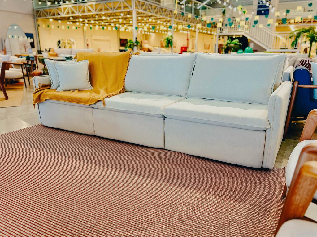 Sofá Retrátil Cama Dinamus Claro | Ciello Sofá Dinamus de 3 lugares na cor clara, com encosto de almofadas soltas e manta decorativa mostarda, ambientado fechado em um showroom iluminado.