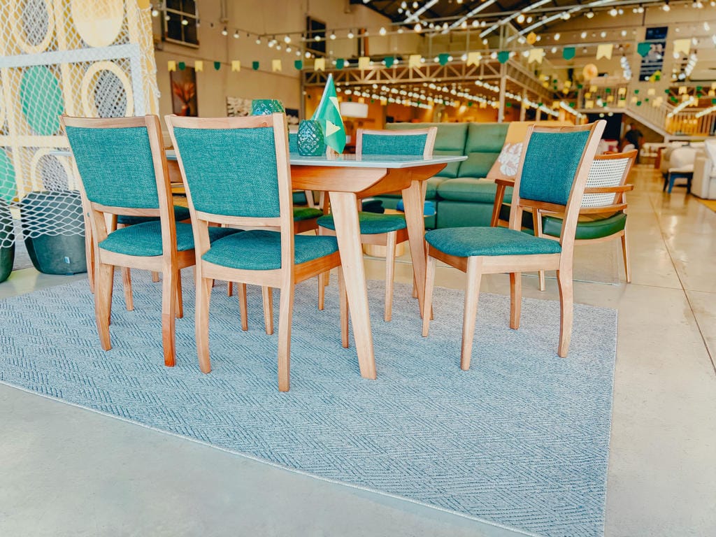 sala de jantar sofia copa do mundo 01 Teal-upholstered wooden dining chairs around a mid-century table on a blue rug in a bright showroom display.