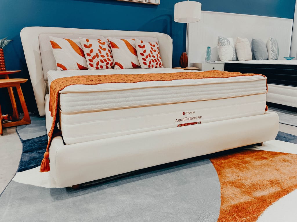 Colchão Aspen Conforto Casal Cama Estofada Colchão Aspen Conforto de casal sobre cama estofada clara, decorado com manta e almofadas alaranjadas, inserido num quarto com parede azul escuro e tapete geométrico.
