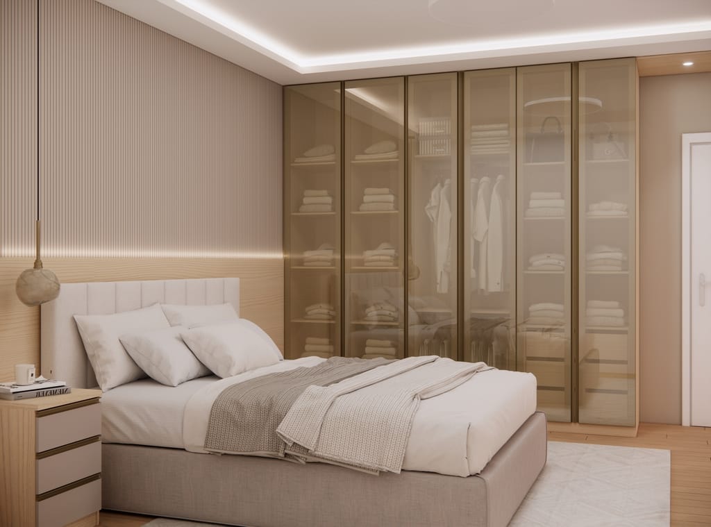 Quarto de casal requintado com uma cama estofada clara encostada a um painel de parede amadeirado e texturizado. À direita, um grande guarda-roupa apresenta portas de vidro reflecta bronze com perfis escuros e iluminação interna em LED, deixando transparecer a arrumação de roupas, prateleiras e cabideiros.