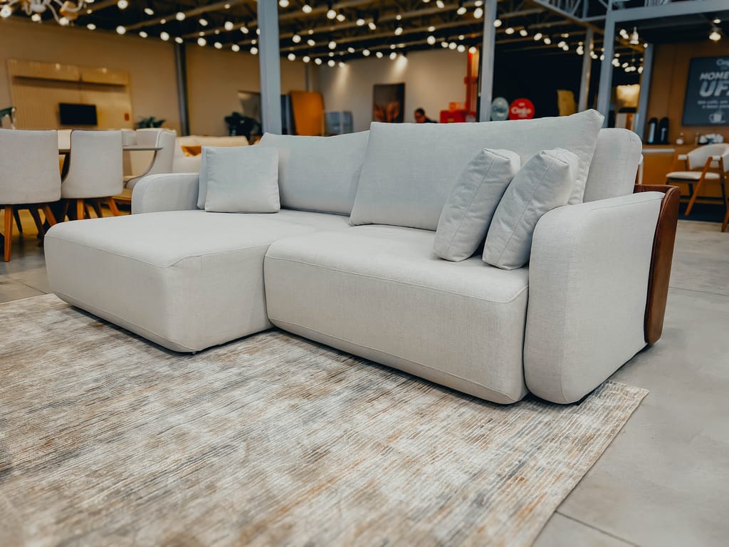 Sofá Retrátil Greevile Off-White Sala TV Sofá retrátil Greevile off-white visto de frente e parcialmente aberto, sobre um tapete claro em um showroom de móveis.